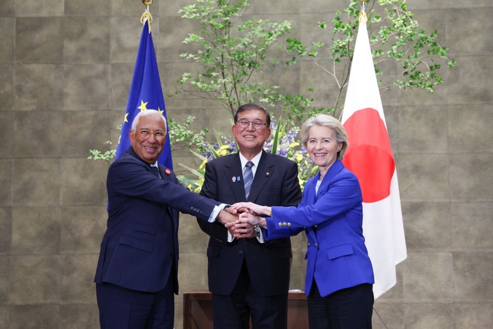 António Costa, Shigeru Ishiba and Ursula von der Leyen shake hands. Behind them is a flower arrangement, flanked to the left by the EU flag and to the right by the Japanese flag.