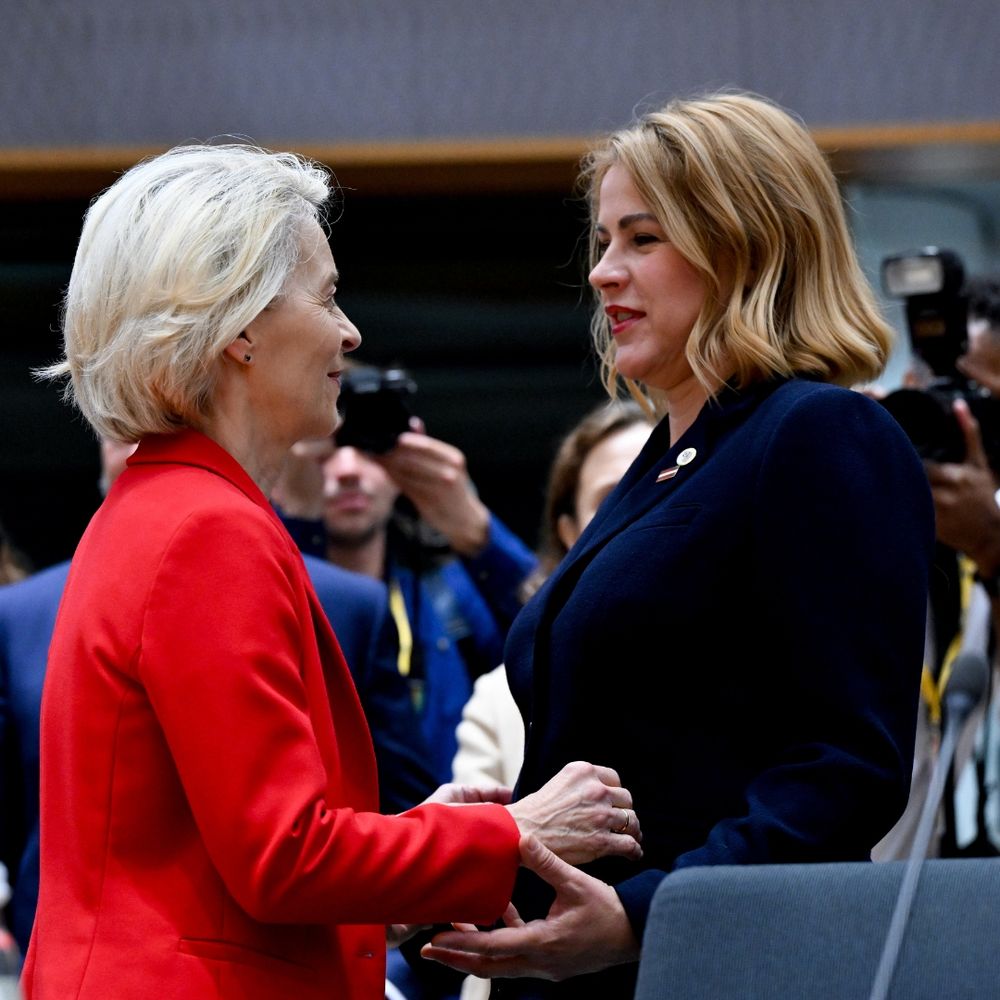 A woman in a red suit (European Commission president) greeting another woman with a blue suit. They are both blond and smiling. Behind them a few cameras.