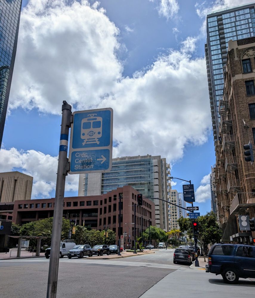 View of Broadway in San Diego looking west with a sky blue colored trolley station wayfinding signage in foreground and bright blue sky with cumulus clouds in background