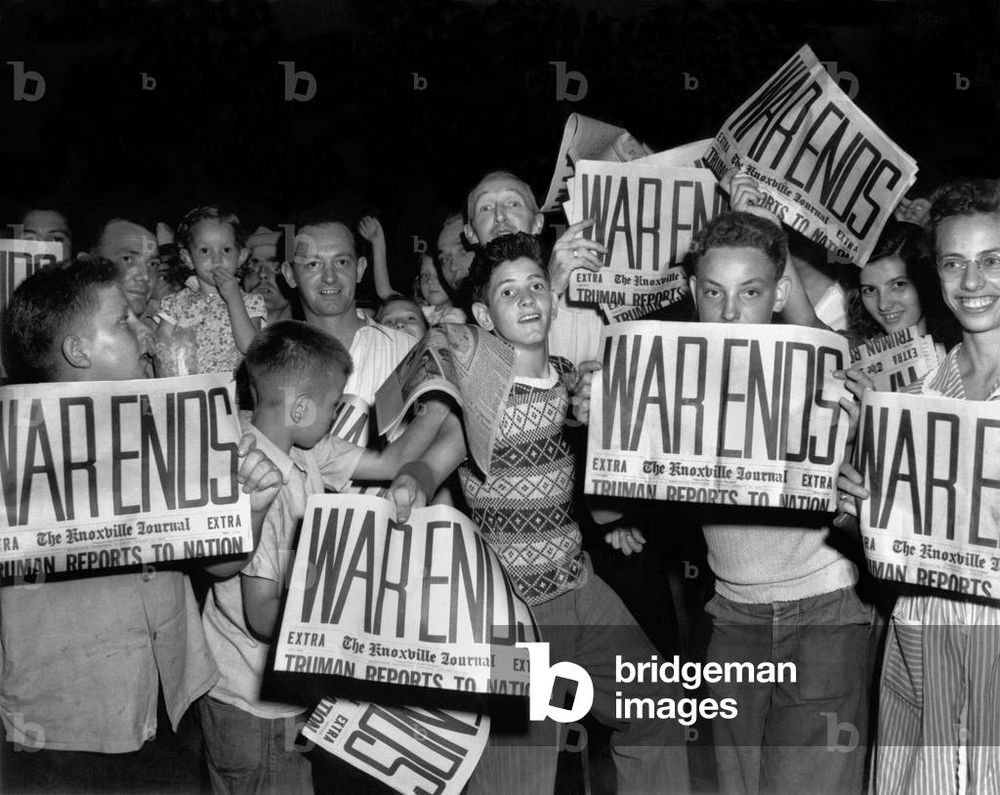 A black and white photo of a group of children crowded together on a street in Tennessee smiling and holding up newspapers headlined 'war ends'