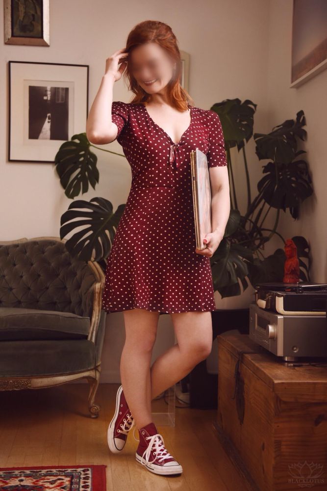 Audrey wears a short maroon dress with white polka dots and a small bow resting against her cleavage. She wears matching maroon converse sneakers, and she carries a stack of vinyl records while standing next to a record player. 