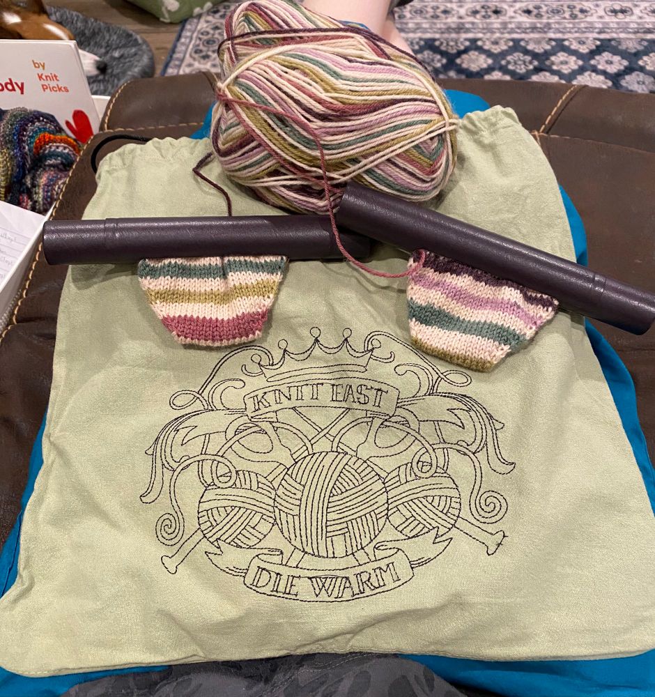 A sage green project bag reads "Knit Fast, Die Warm" in black embroidery with a design of scissors and yarn balls and knitting needles and a crown. On top of the bag are two socks-in-progress. Currently they are just stripey toes disappearing into dark browny purple tubes. The socks are cream, sage, forest, raspberry, mauve, and brown stripes. Two small striped balls of yarn sit above them.
#knitting #knit yarn ball 🧶 