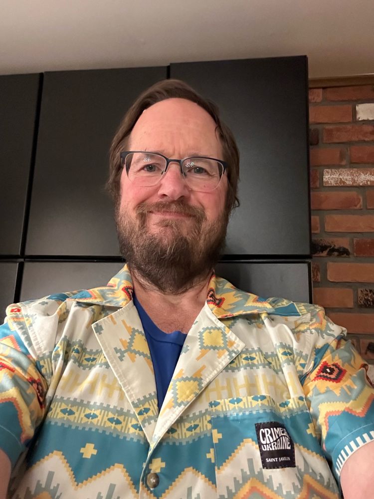 An old bearded white man stands in front of a pantry wearing a blue and yellow Hawaiian style shirt with a Ukrainian pattern on it.