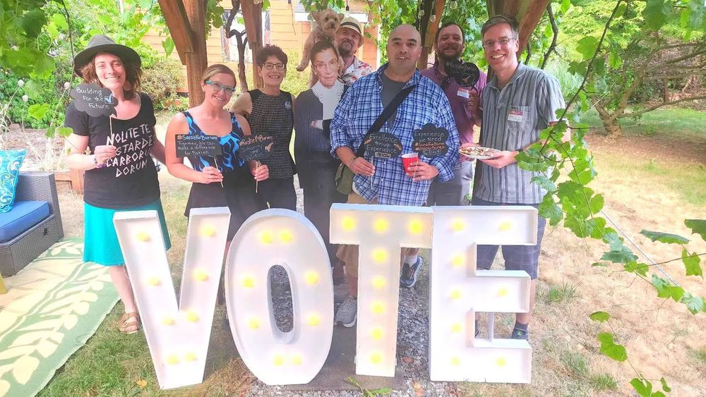 Ginele, Rashell, Sarah, Daniel, Taco Charlie (a small dog), Hugo, Rocco, and Sam stand with a cardboard cutout of Ruth Bader Ginsberg behind giant white letters that spell out VOTE