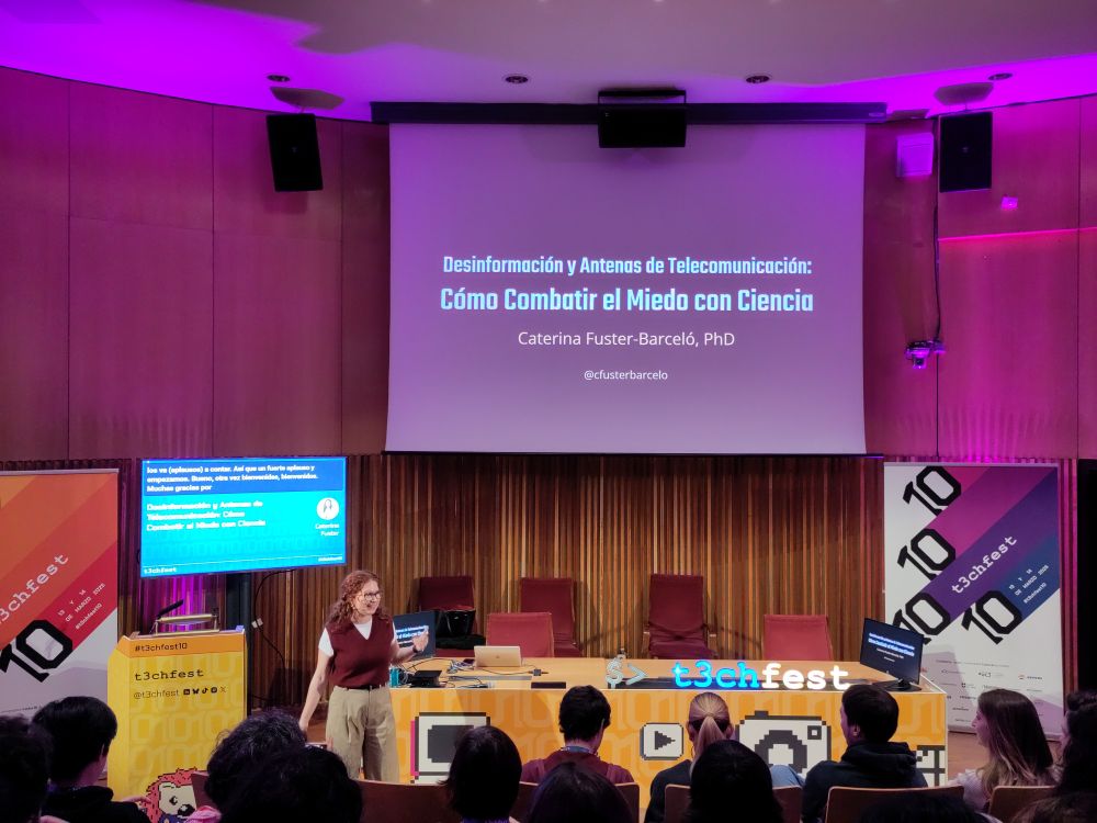 Foto de una mujer en un escenario, en la pantalla gigante se lee "Desinformación y antenas de telecomunicación: Cómo combatir el miedo con ciencia"