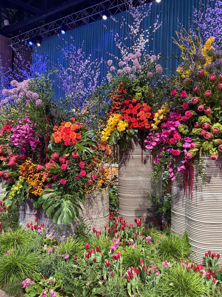 A vibrant indoor floral display featuring tall cylindrical planters overflowing with colorful blooms including roses, orchids, and tulips. The arrangement is set against a backdrop of teal curtains and twinkling lights, with flowering branches rising above the main installation and tulips and pink flowers planted below.
