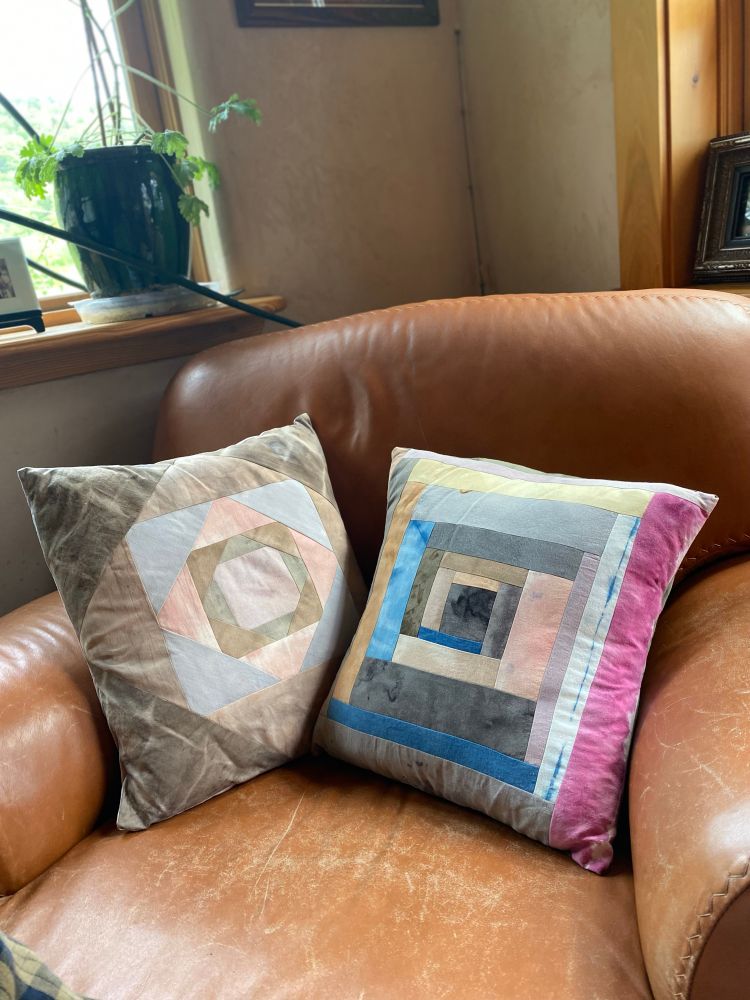 Two more patchwork pillows on the same brown leather chair. The one on the left is made with lavender, green, pink, light blue, and different shades of brown in a pattern that begins with a square in the middle and adds four triangles, one on each side, again and again to get a bigger square. The pillow on the right is made from strips of fabric in a log cabin pattern, kind of like a square spiral. The strips are in different shades of grays, blues, pinks, yellows, with some lavender, green, and brown. 