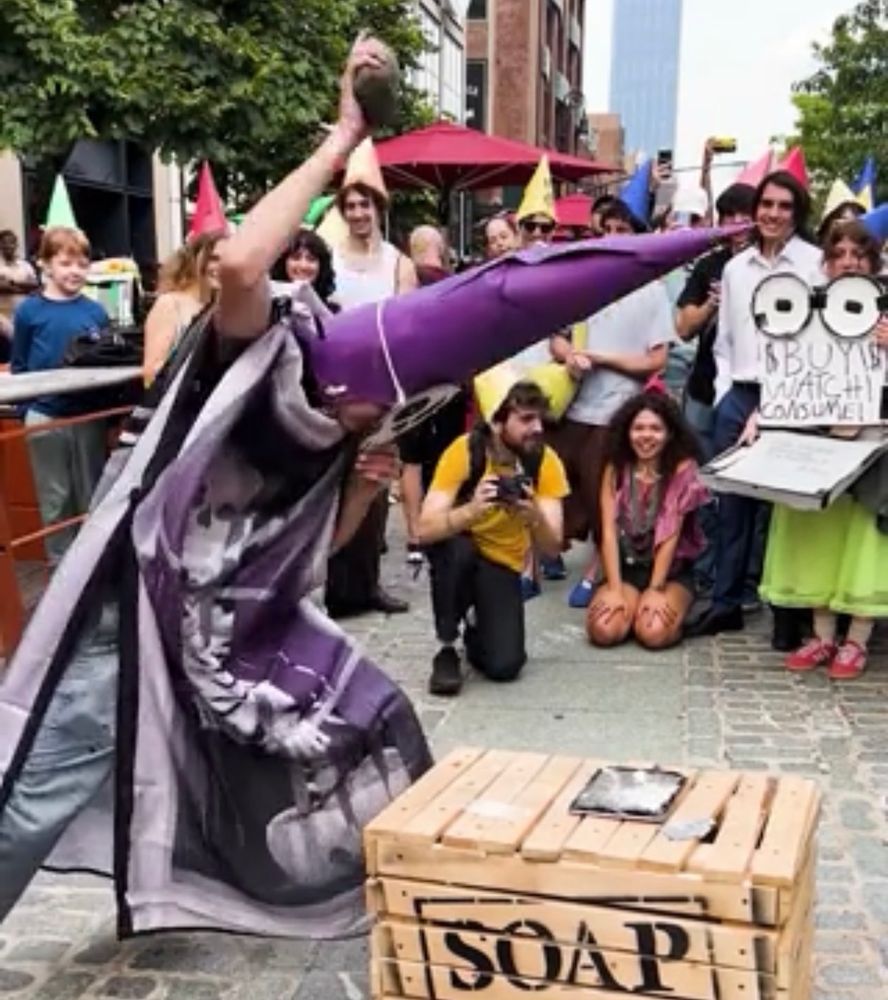 A man in a purple robe and very tall, pointy hat … well, it extends down the back of his head, so maybe more of a hood … and a skull mask, smashing an iPad with a rock.