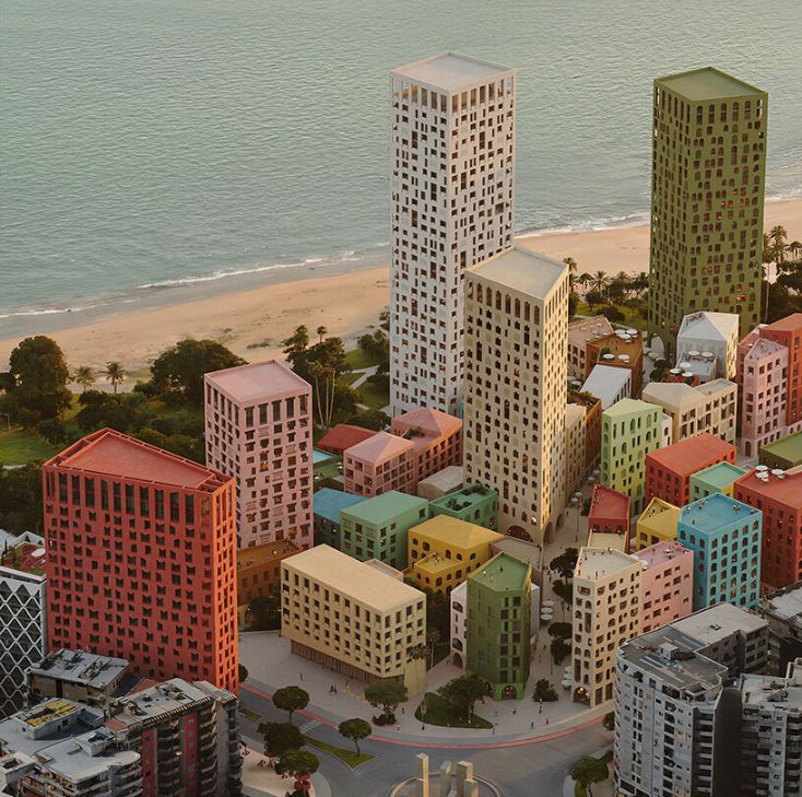 Aerial view of a coastal cityscape featuring numerous mid-rise and high-rise buildings in a variety of pastel colors, including red, green, yellow, blue, and beige. The buildings are clustered together near a sandy beach, with the ocean in the background and some palm trees and greenery along the shoreline. The urban layout appears planned and geometric, with curved streets and small plazas interspersed among the buildings.