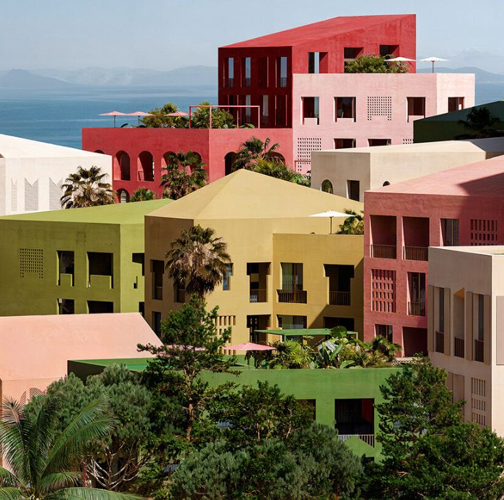 Aerial view of a coastal cityscape featuring numerous mid-rise and high-rise buildings in a variety of pastel colors, including red, green, yellow, blue, and beige. The buildings are clustered together near a sandy beach, with the ocean in the background and some palm trees and greenery along the shoreline. The urban layout appears planned and geometric, with curved streets and small plazas interspersed among the buildings.