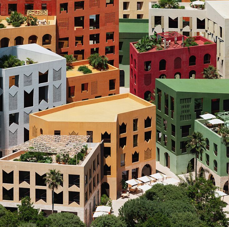 Aerial view of a coastal cityscape featuring numerous mid-rise and high-rise buildings in a variety of pastel colors, including red, green, yellow, blue, and beige. The buildings are clustered together near a sandy beach, with the ocean in the background and some palm trees and greenery along the shoreline. The urban layout appears planned and geometric, with curved streets and small plazas interspersed among the buildings.