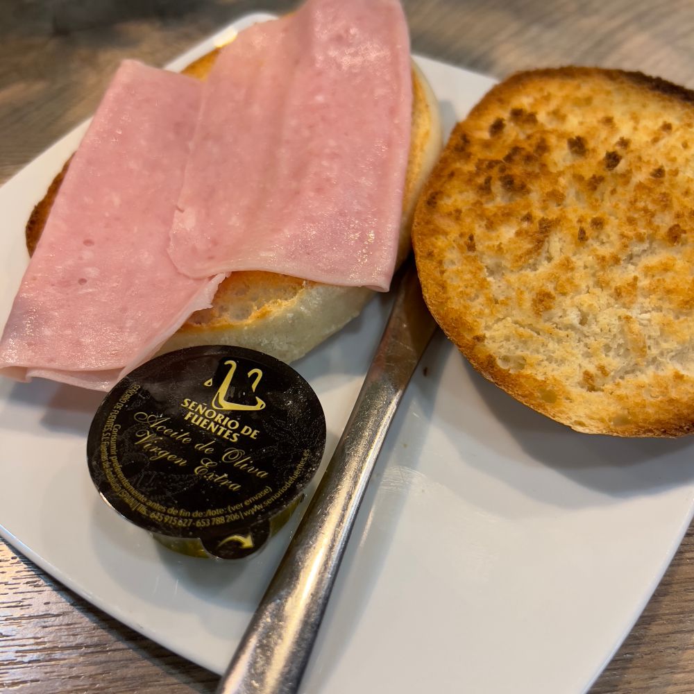 En la imagen se puede ver un plato de forma parecida a un cuadrado con los lados ligeramente curvos, y de color blanco. Sobre el plato hay dos medias tostadas: una de las mitades tiene un par de lonchas de jamón cocido. En el plato también hay un cuchillo de untar y un pequeño envase de aceite de oliva virgen extra.