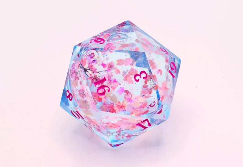 Clear light blue overside D20 dice with pink numbers and cherry blossons decorating it, it has a liquid core filled with petals and glitter