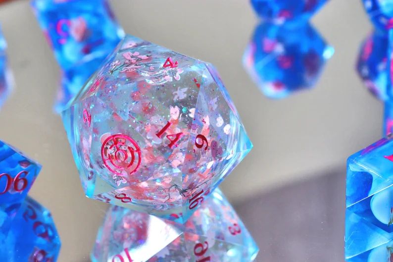 Clear light blue overside D20 dice with pink numbers and cherry blossons decorating it, it has a liquid core filled with petals and glitter