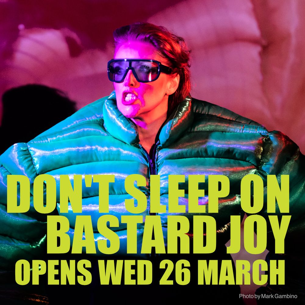 Fierce looking redheaded woman in an oversized green puffer jacket giving electro-clash attitude, wearing matching green club sunnies. 