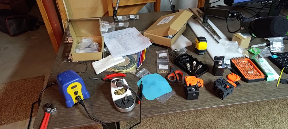 A picture of a workbench surface, cluttered in various 3D printer parts, a soldering iron, several screwdrivers, a wrench, and a microphone and keyboard.