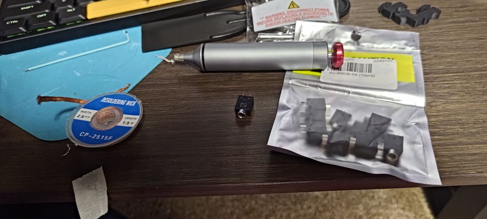 A photo of a desk containing a broken headphone jack, a bag containing a bunch of replacements, a solder sucker and desoldering wick.