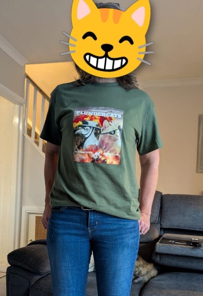 A woman wearing a Plundercats the movie T shirt. There is a grinning cat emoji over her face. The back end of a tortie cat can be seen on the sofa behind her.
