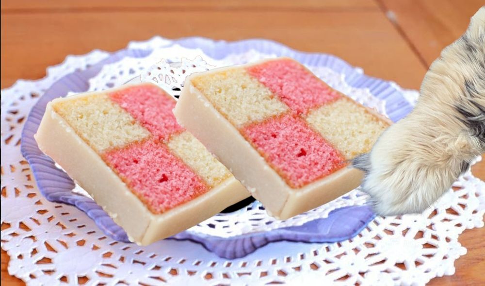 Two slices of Battenberg cake on a doily covered plate. A tortie cat paw is pushing the plate