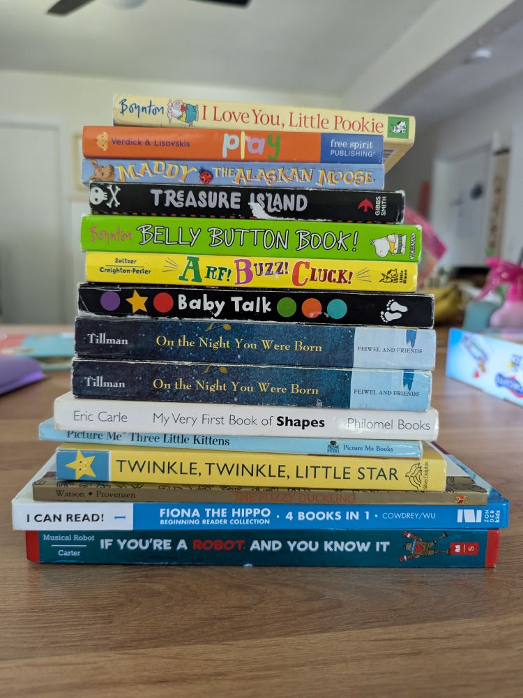 A moderately high stack of fifteen children's books sits on a butcher block counter. Some of the titles displayed are: "I Love You, Little Pookie", "Play", "Maddy the Alaskan Moose", "Treasure Island", "Belly Button Book!", "Arf! Buzz! Cluck!", "Baby Talk," two copies of "On the Night you Were Born," "My Very First Book of Shapes," "Picture Me Three Little Kittens", "Twinkle Twinkle Little Star", "Fiona the Hippo" and "If You're a Robot and You Know It."