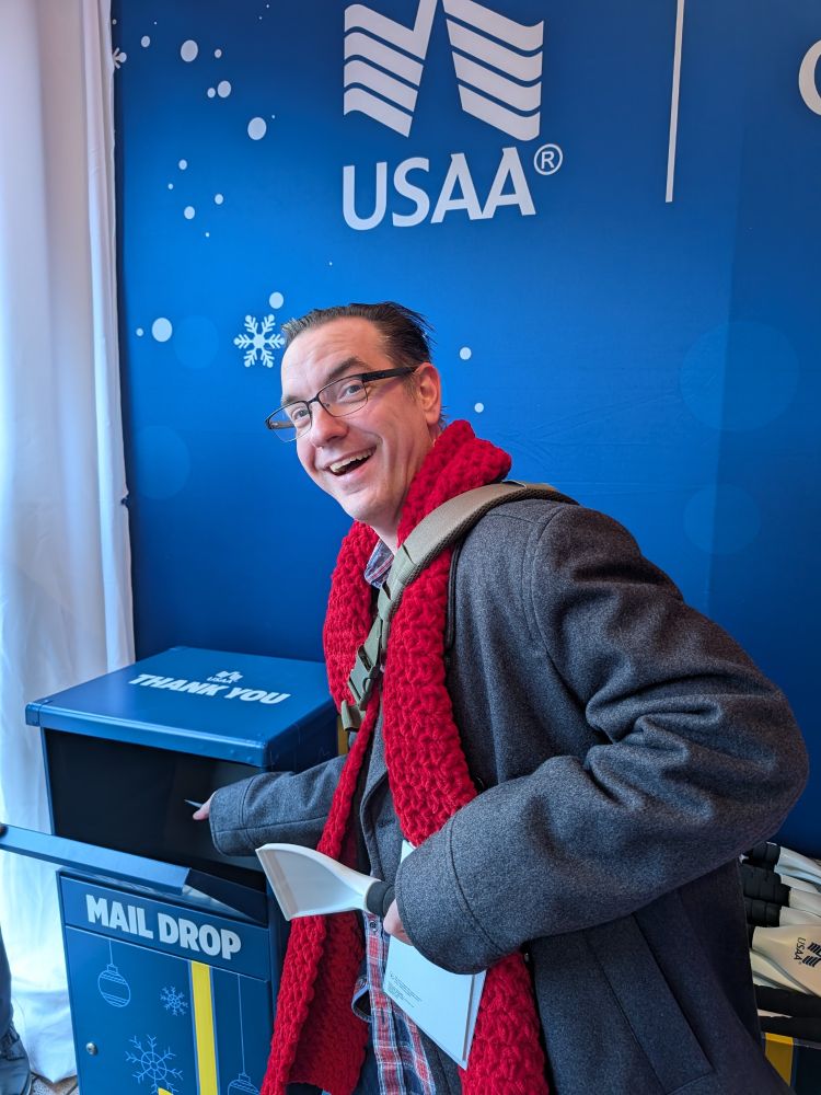A middle aged white guy with a USAA ice scraper deposits a postcard in a USAA mail box, for troops