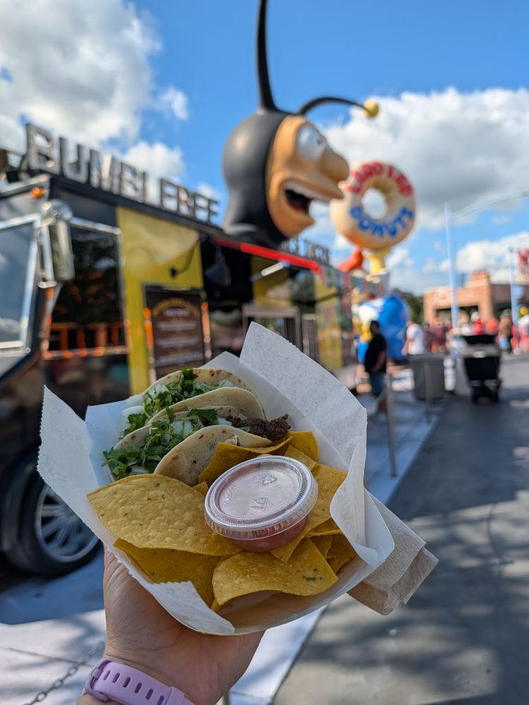 A hand holds a paper tray of carne asada tacos and tortilla chips with a plastic cup of salsa, in front of Bumblebee Man's Taco Truck in Universal Studios (Orlando)