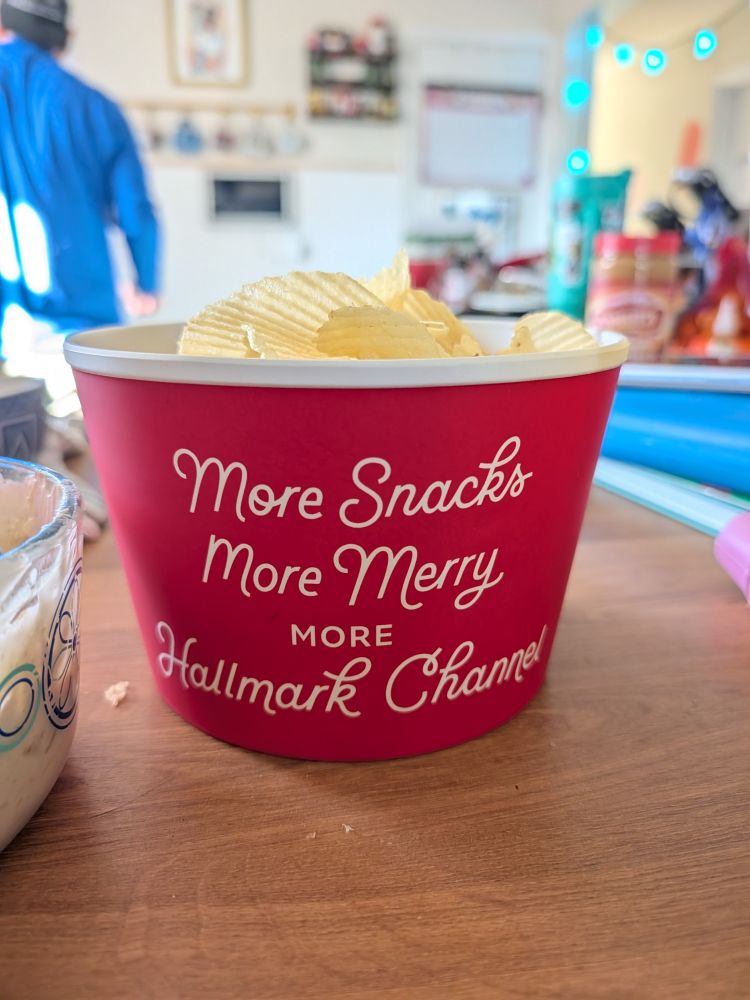 A red bowl with the words "more snacks, more merry, more Hallmark Channel" sitting on a kitchen counter, containing Ruffle potato chips 