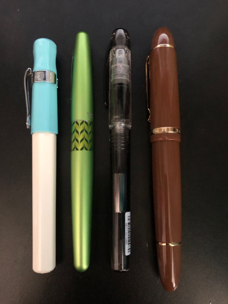 Four fountain pens laid side by side. From left to right: Blue Pilot Kakuno (With a Kaweco clip attached to the cap), Pilot Metropolitan in green, Platinum Preppy in black, and Jinhao x159 in brown.