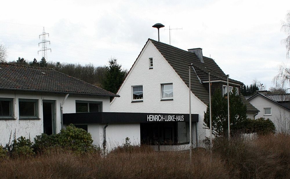 Ein weißes Einfamilienhaus mit Anbau am Waldrand, auf dem Dach eine Luftschutzsirene.