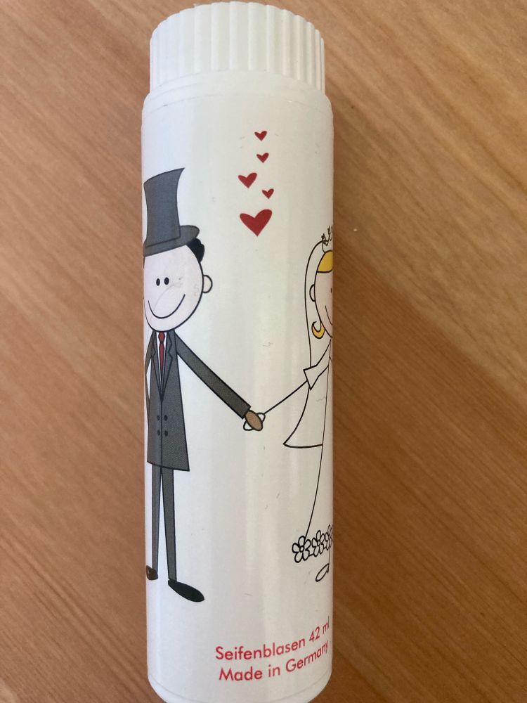 Weiße Seifenblasendose mit darauf gezeichnetem Bräutigam und Braut, die sich an der Hand halten. Dazwischen rote Herzchen.
Unten steht in roter Schrift: Seifenblasen.