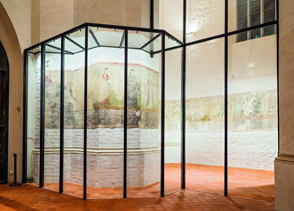 Ausschnitt des originalen Totentanzfreskos, schemenhaft sind bunte Figuren zu erkennen. Das Fresko an der Wand der Turmhalle ist von einer Glaswand geschützt und hell erleuchtet.