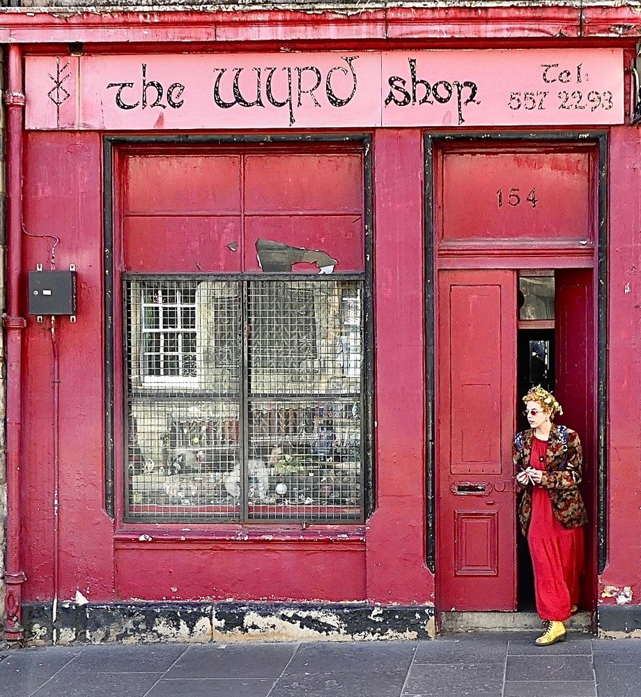 A woman dressed in red emerges from the red frontage of The Wyrd Shop, keys in hand.