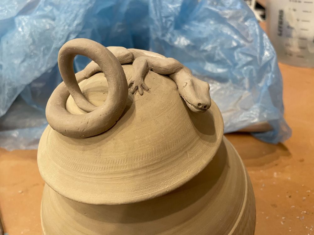 A photo of an unfired clay jar about 8 inches high and 6 around with an abstract texture around the middle and a domed lid. On the lid is a cute little lizard wrapped around the dome, peering forward at you. Its tail curls up into a round handle for the lid.