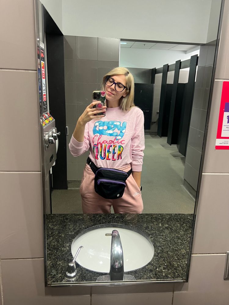 Christie smiles in the mirror of an airport bathroom. She is wearing a pink long sleeved tee that says “cozy chaotic queer” on the front. 