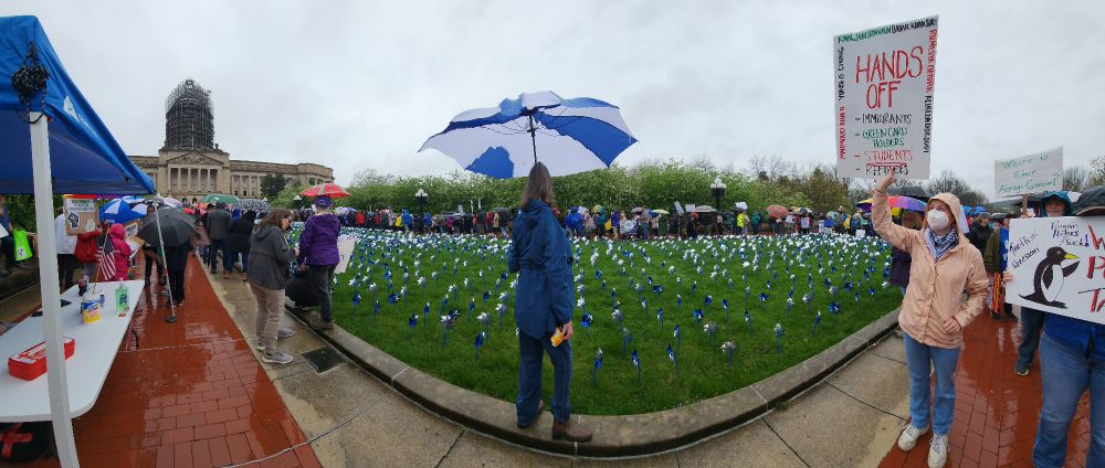 Panorama shot of myriad protestors standing on either side of the lawn in front of the Kentucky Capitol on a gray and rainy day. One person's sign reads, "HANDS OFF -IMMIGRANTS-GREEN CARD HOLDERS -STUDENTS -REFUGEES" and lists the names of people unjustly and illegally apprehended recently by ICE, e.g., "RUMEZYA OZTURK".