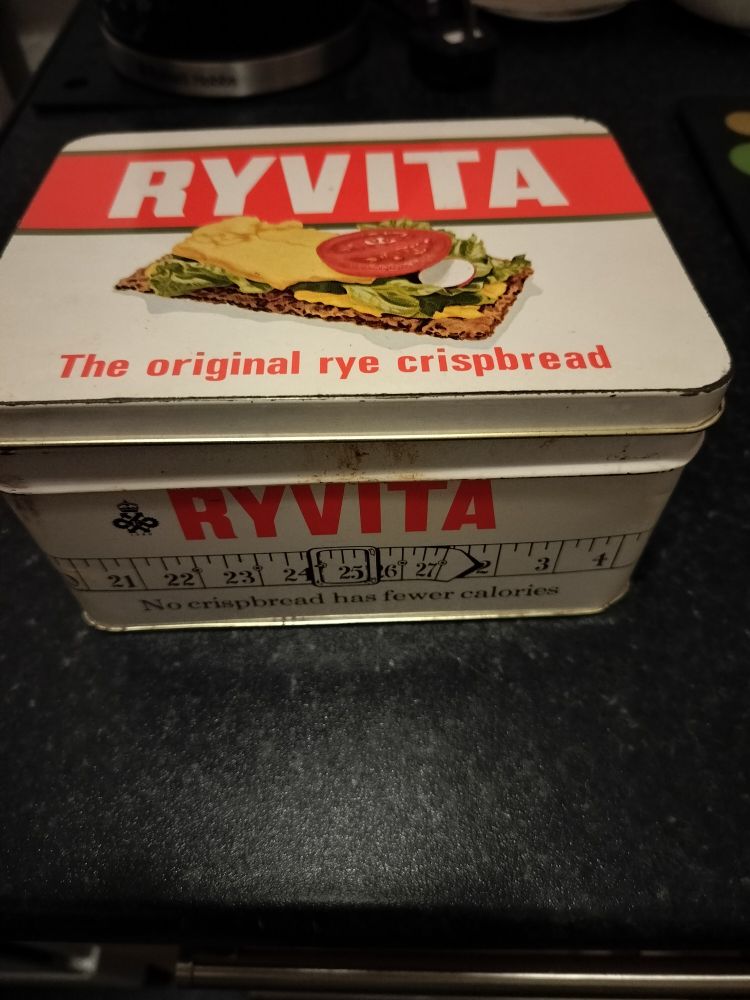 Retro style Ryvita crispbread tin with image of a "tasty" looking cracker loaded up with cheese & salad. There's also an image of a tape measure wrapped around the tin showing the "desired" 25 inch waist measurement that encouraged desperate dieters at the time to eat said cardboard texture crackers. 😑 Disdainful emoji face.