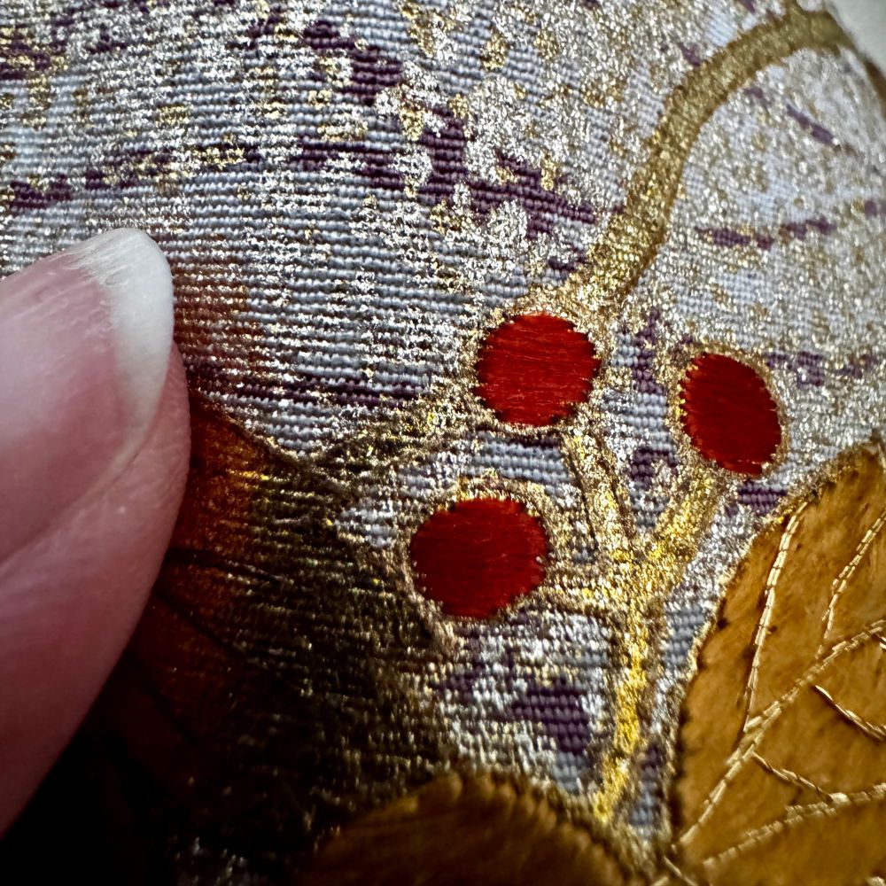 A hand holds up kimono fabric with delicate red embroidered berries, coppery embroidered leaves, with fine gold cord for the leaf veins on top of the painted floral scene and sweeping background fabric of white and grey with soft edges mottled by touches of golden paint.