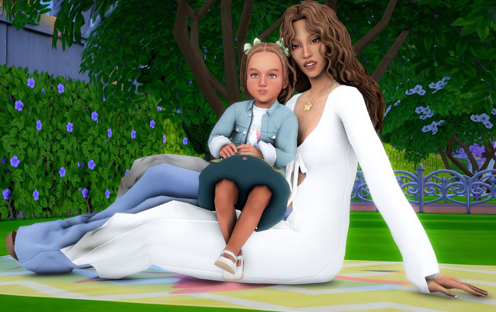 A toddler Ophelia sits in the lap of her mother Evelyn. They are sitting on a picnic blanket in a park