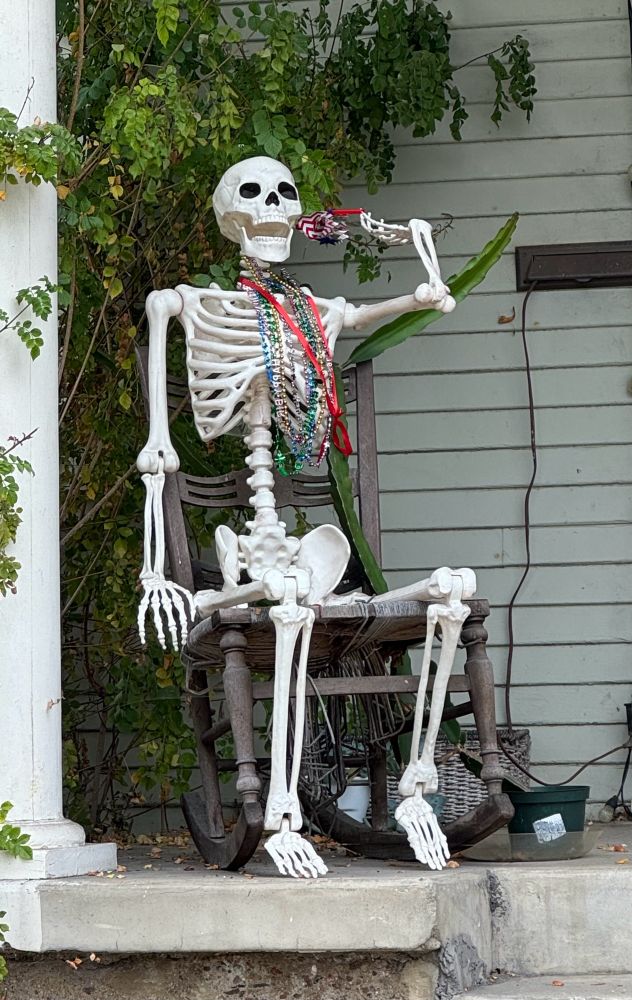 Human skeleton wearing Mardi Gras beads relaxes comfortably in wooden chair on front porch