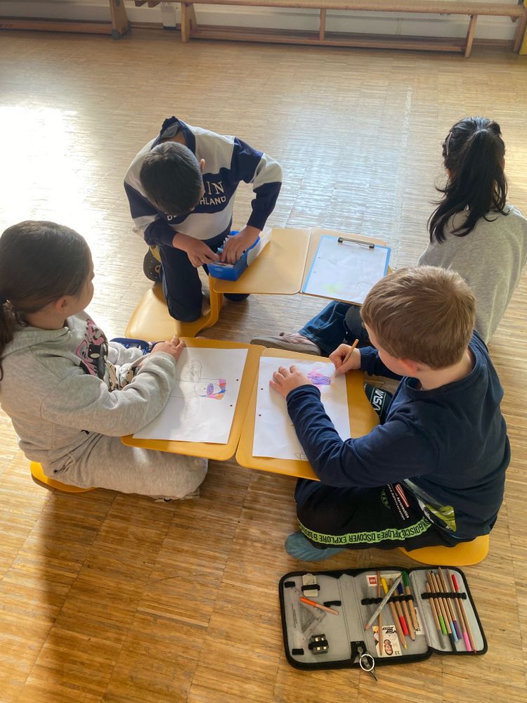 4 Kinder sitzen auf dem Boden, jedes Kind hat eine Tisch-Stuhl-Kombination, es sitzt auf einer Fläche, verbunden damit ist eine Schreibfläche.
