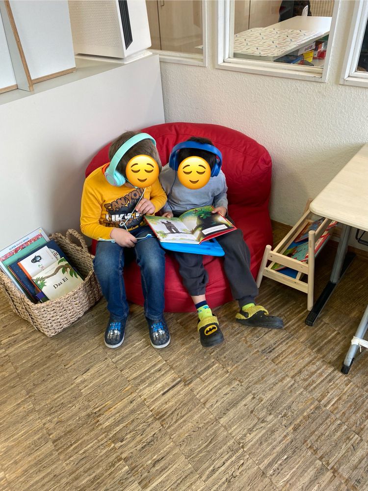 2 Kinder sitzen auf einem Sitzsacksofa und hören über Kopfhörer, lesen dabei in einem Buch mit.
