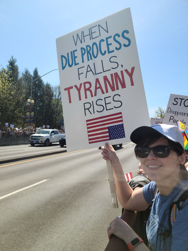 The poster, a young white woman with short brown hair, standing along the side of a bridge with traffic going by. She is wearing a white baseball cap and sunglasses and holding a protest sign that reads, “When due process falls, tyranny rises” with an upside down U.S. flag at the bottom. 