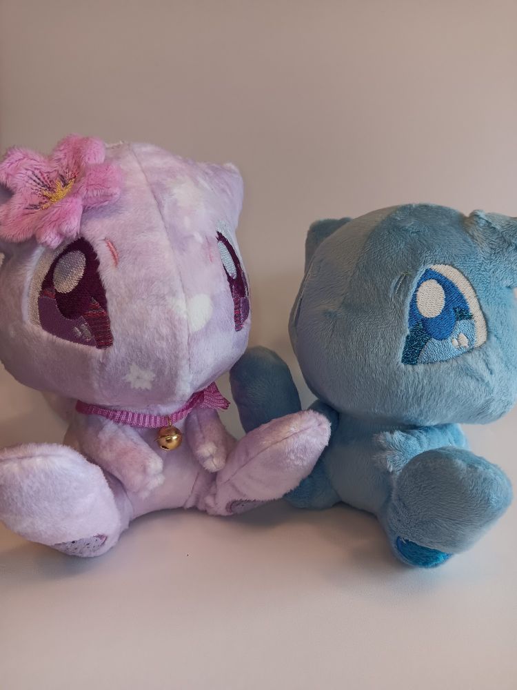 Two of the same kind mew plushies. The shiny one as in the previous picture, same background, is sitting next to a Light pink one, which also has cherry blossoms printed on. It wears a pink Band around its neck with a bell and a big cherry blossom as accesoire at its ear.