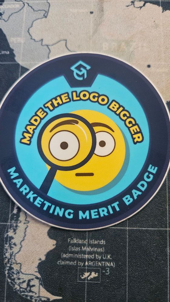 Round sticker with the words Make the Logo Bigger Marketing Merit Badge. The interior has a smiling face emoji with a magnifying glass.