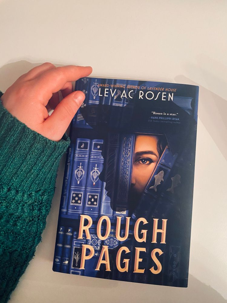 A hardcover copy of Lev AC Rosen's Rough Pages is placed on a white table. My white, left hand in a dark green sleeve is framing the book to the left.