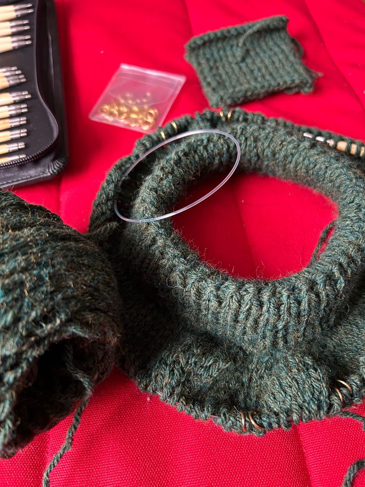 photo of several knitting items on a red chair. starting at 11 going clockwise, a set of bamboo knitting needles. a pack of gold stitch markers. a gauge swatch knit in stockinette. a knit sweater project on circular needles. the sweater is being knit top down (collar first). the collar is k1p1 ribbing folded over and the body is stockinette. the green yarn being used to knit the sweater.