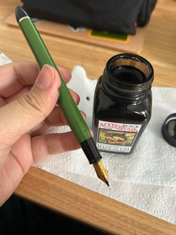 hand holding a green fountain pen. behind it is a jar of open noodlers black ink placed on top of toilet paper to absorb spills. behind the ink is a sketchbook and pencil case