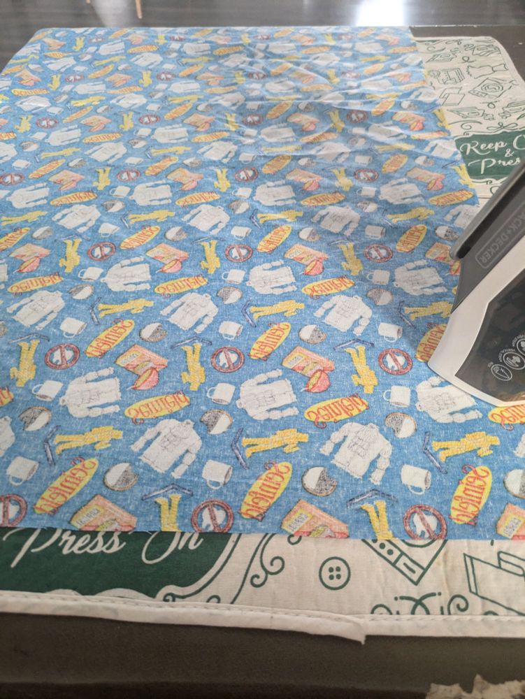 The backside of a piece of Seinfeld-branded fabric, sitting on a pressing mat, next to an iron. 