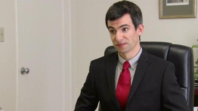 A imagem mostra o humorista Nathan Fielder sentado em uma cadeira de escritório. Ele está vestindo um terno preto com uma camisa cinza e uma gravata vermelha. Ele tem cabelo castanho escuro e está olhando para a câmera. O fundo é uma parede branca com uma porta e uma pintura.