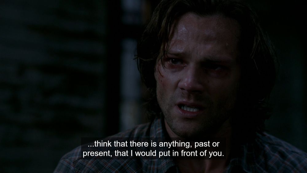 Sam Winchester in a plaid shirt looking visibley upset with subtitles, standing against a dark background. In supernatural 